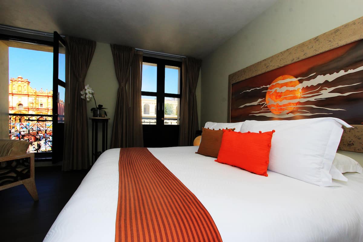 Quarto de hotel com uma cama grande, uma janela com cortinas, um quadro de um pôr do sol e vista para um edifício lá fora.