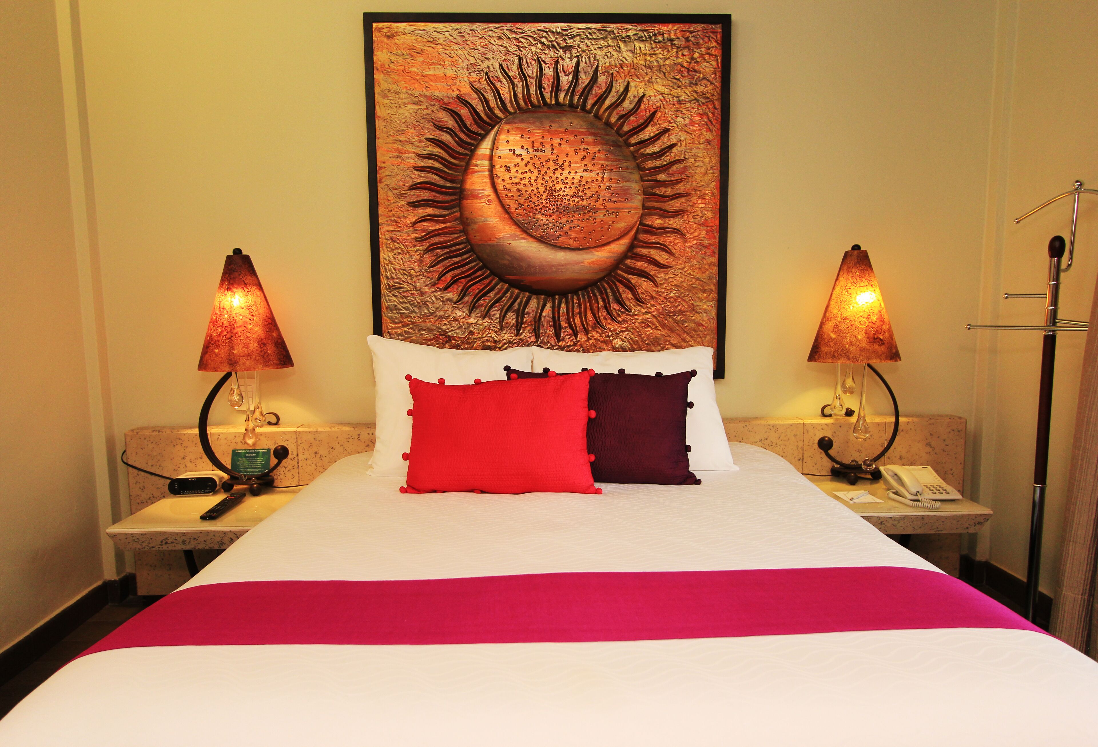 A hotel room with a large bed, two bedside lamps, a telephone, and a painting of a sun on the wall.