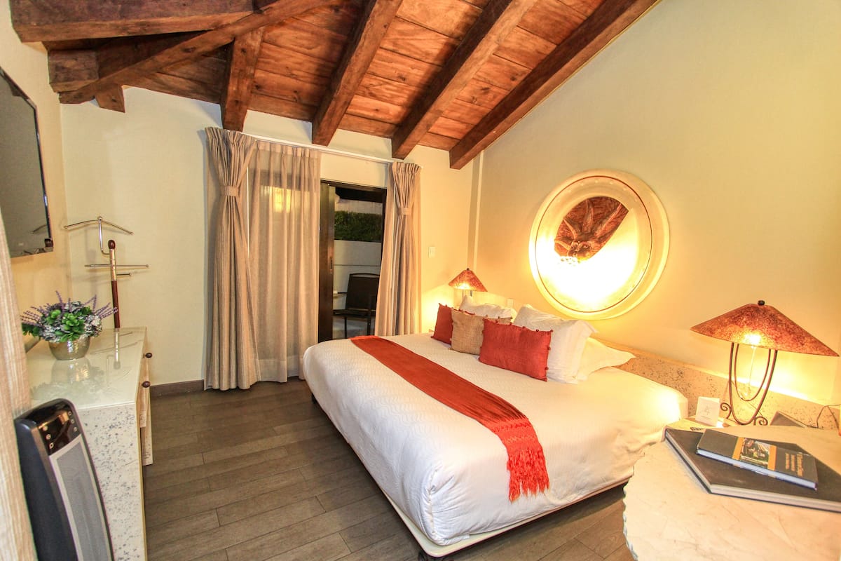 Quarto de hotel com uma cama grande, uma mesa com computador, uma luminária e uma janela com cortinas.