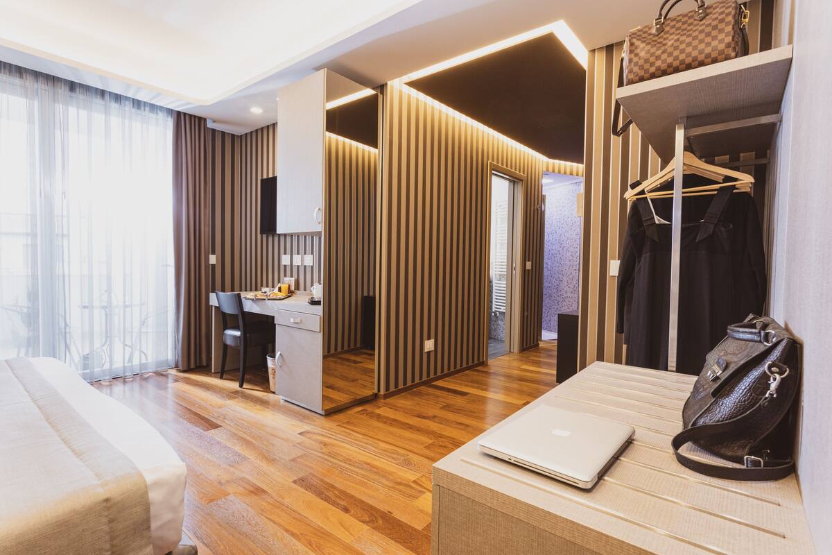 Ein modernes Hotelzimmer mit Holzboden, einem Bett, einem Schreibtisch mit Stuhl, einem Kleiderschrank und einem Garderobenhaken.