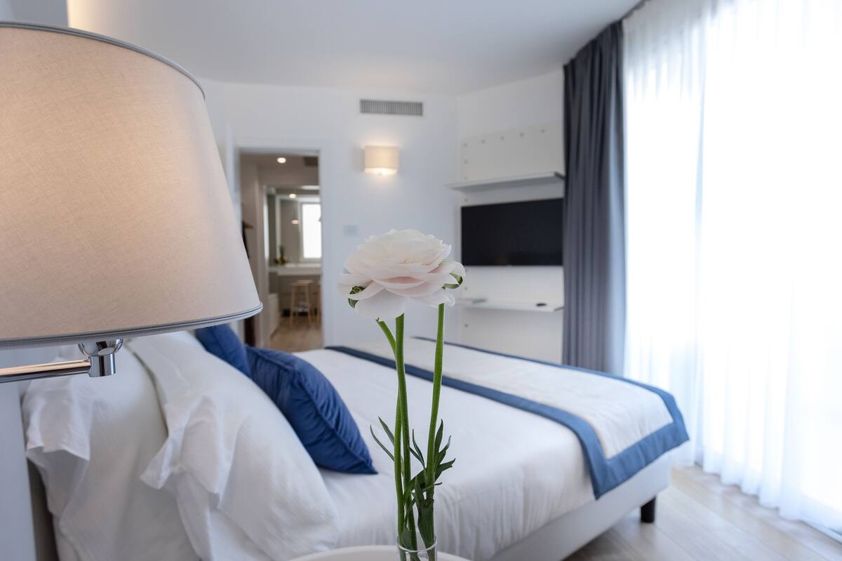 Ein Hotelzimmer mit einem Bett, einem Nachttisch mit Lampe, einer Vase mit einer einzelnen Blume und einem an der Wand befestigten Fernseher.