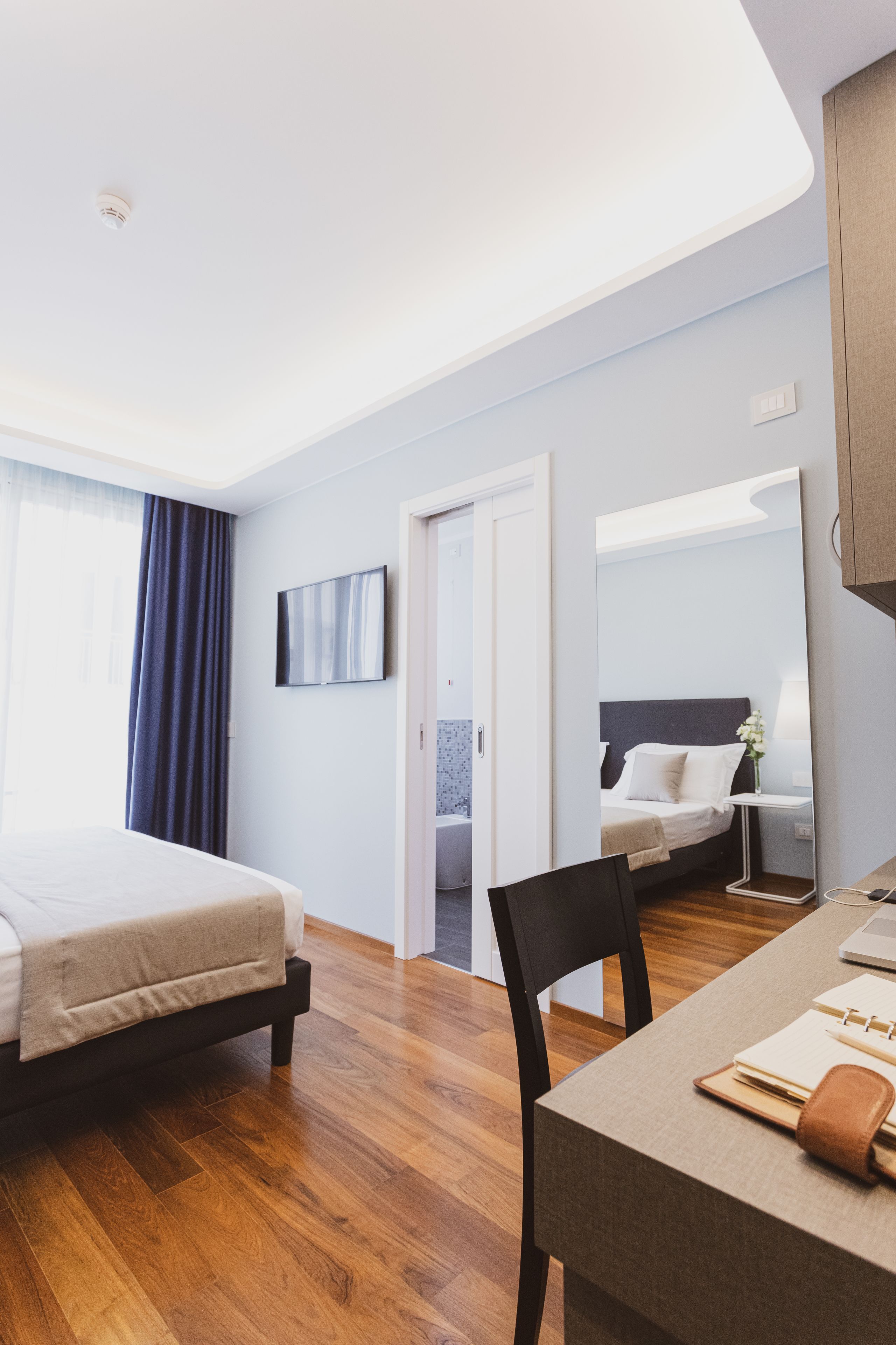 Una habitación de hotel moderna con piso de madera, una cama, un escritorio con una silla, un televisor y un espejo.
