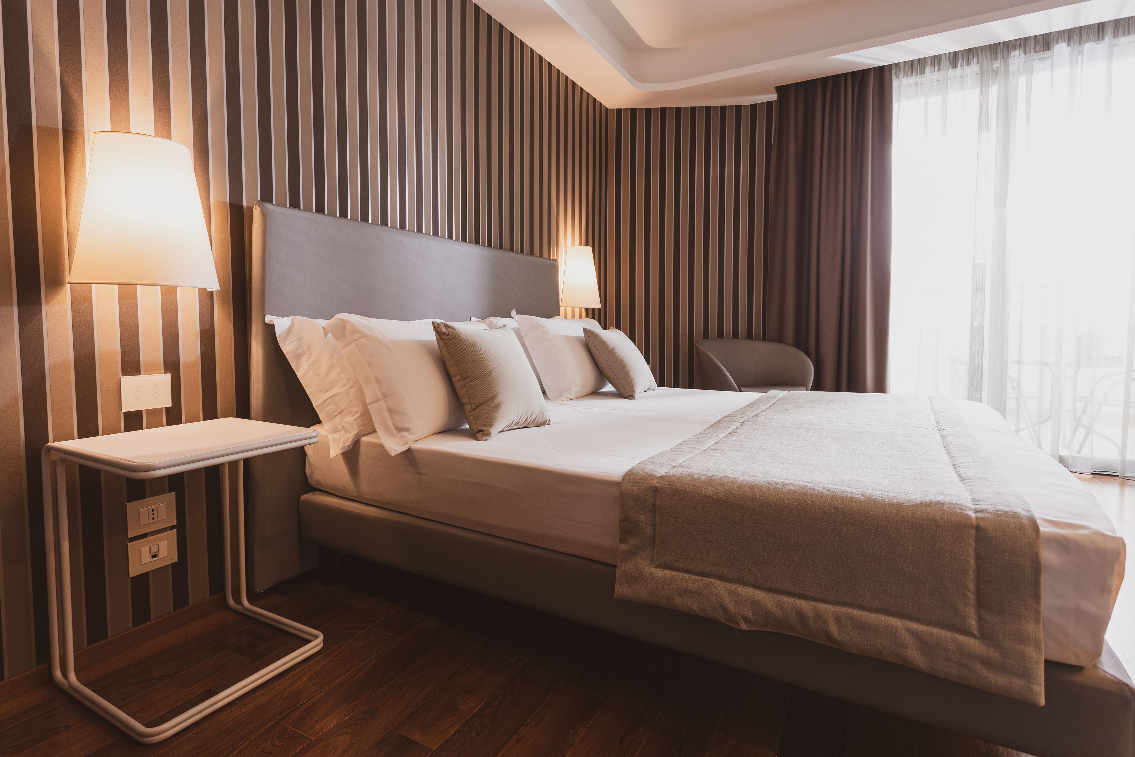 A modern hotel room with a large bed, two bedside tables with lamps, a chair, and a view of the outdoors.