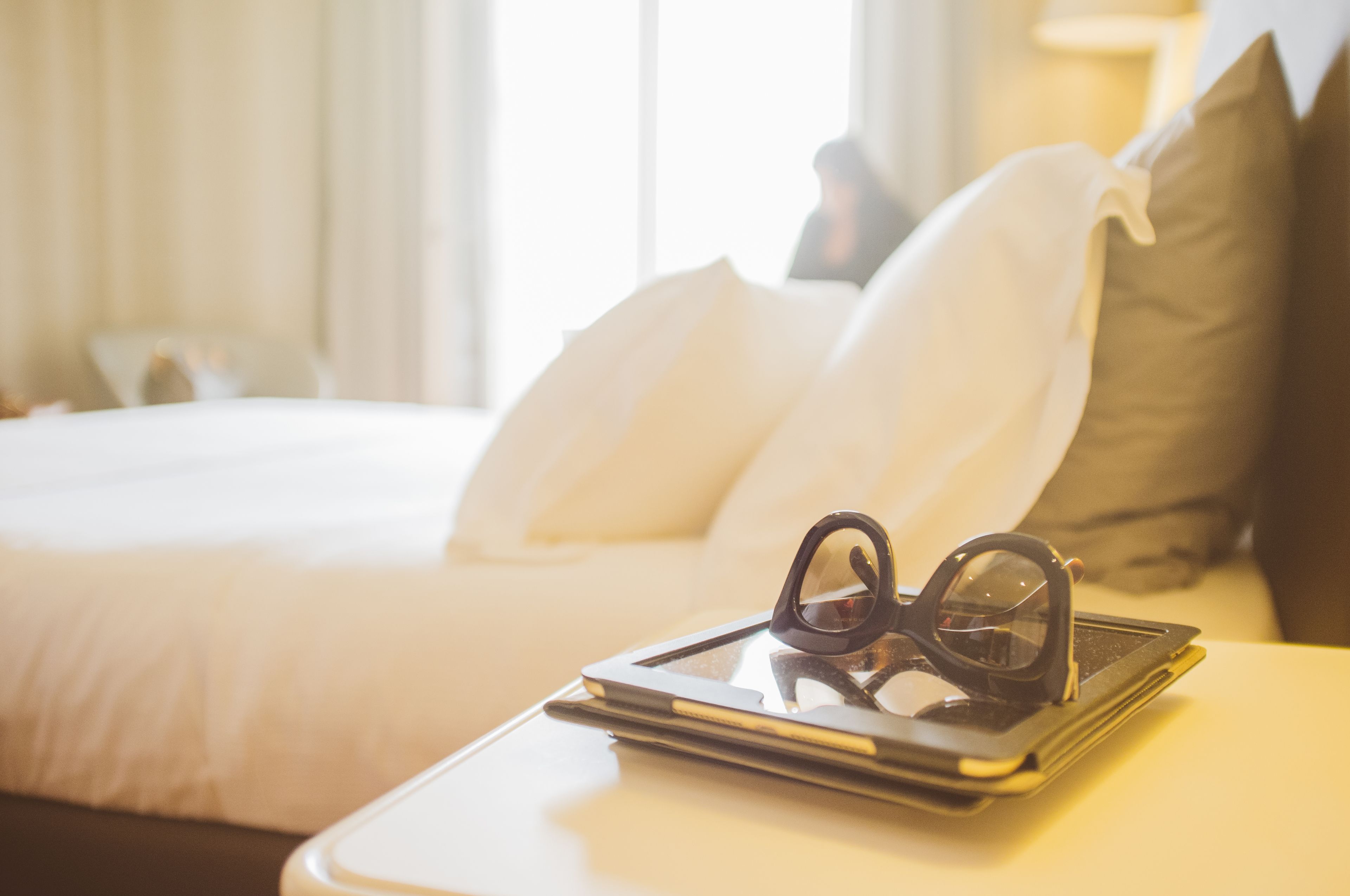 A neatly made bed with white and brown pillows, a tablet with glasses on top, and a bedside table.