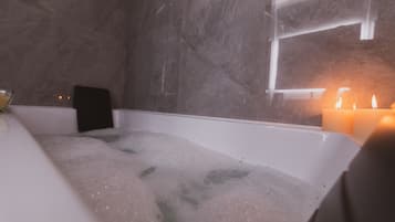 A modern bathroom with a bathtub filled with bubbles, a glass of water, and candles on the side.