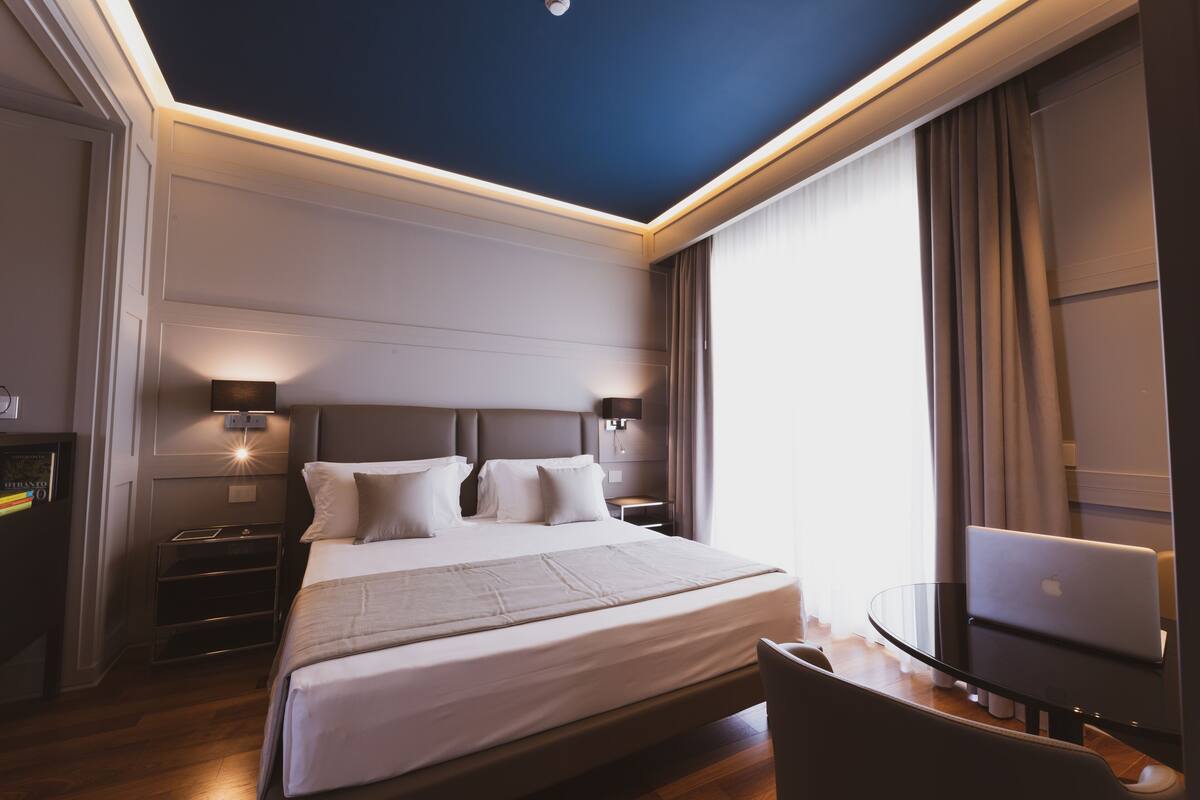 Ein modernes Hotelzimmer mit einem großen Bett, Nachttischen, einem Schreibtisch mit einem Laptop und Blick ins Freie.