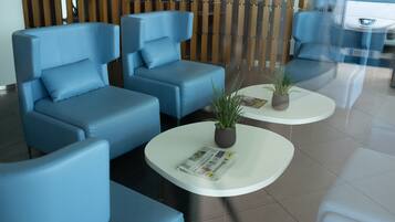 A modern waiting area with blue sofas and white coffee tables, featuring a potted plant and a newspaper.
