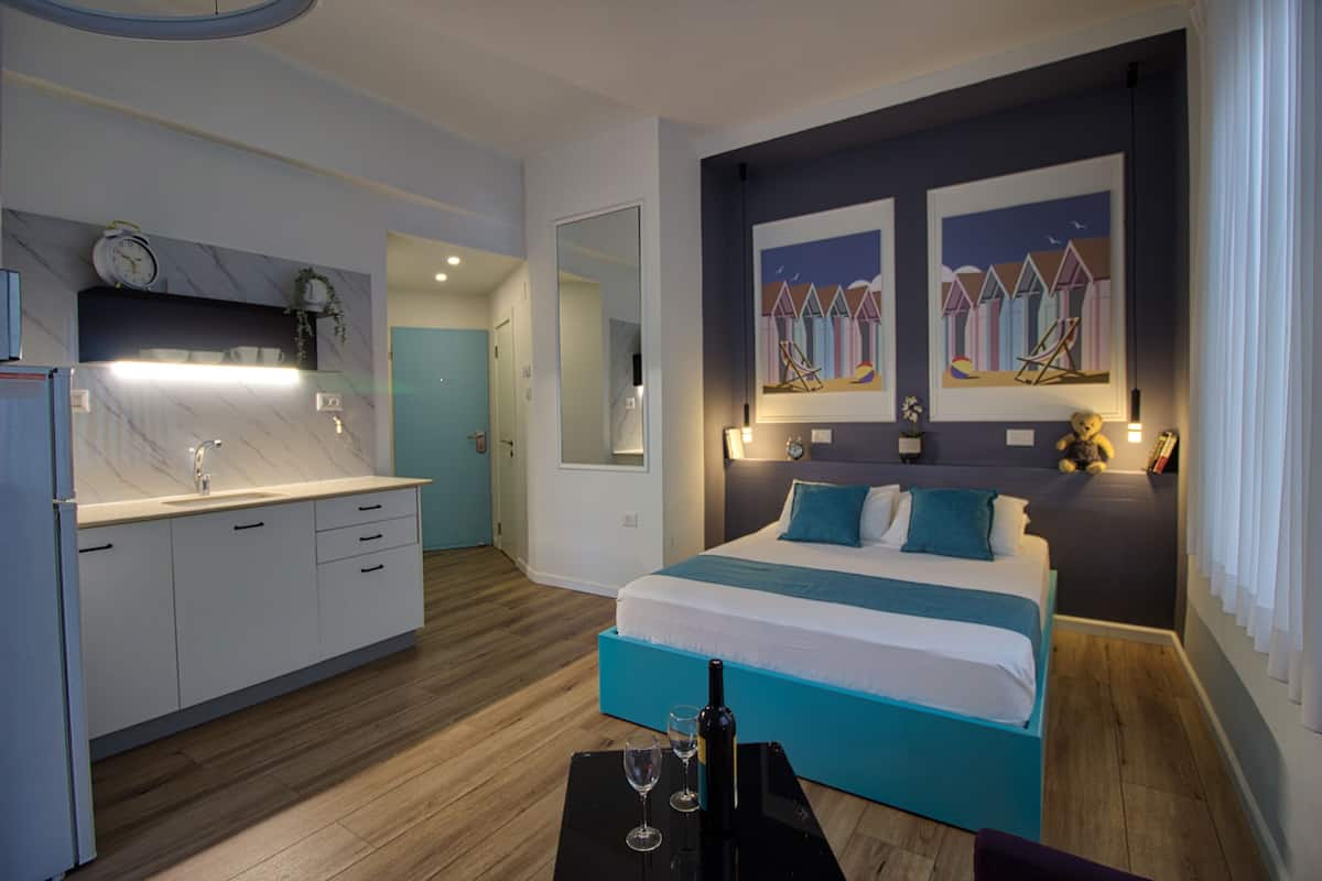 Una habitación de hotel moderna con cama, una pequeña mesa con una botella y un vaso, kitchenette y obras de arte en las paredes.