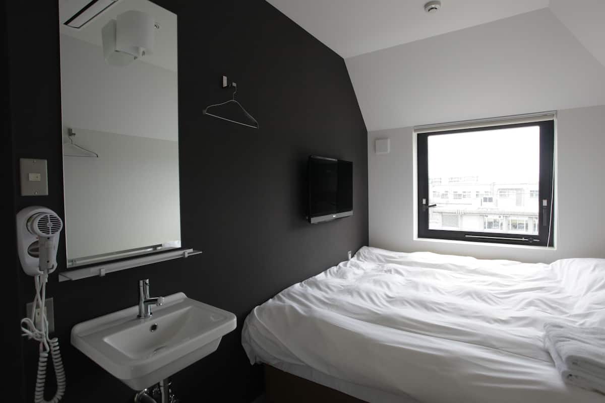 A hotel room with a bed, a white sink, a mirror, a hairdryer, a television, and a window with a view of buildings.