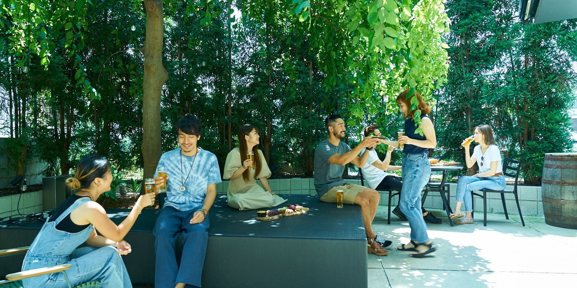 A group of people sitting and standing outdoors, some holding drinks, under trees with a table and chairs nearby.