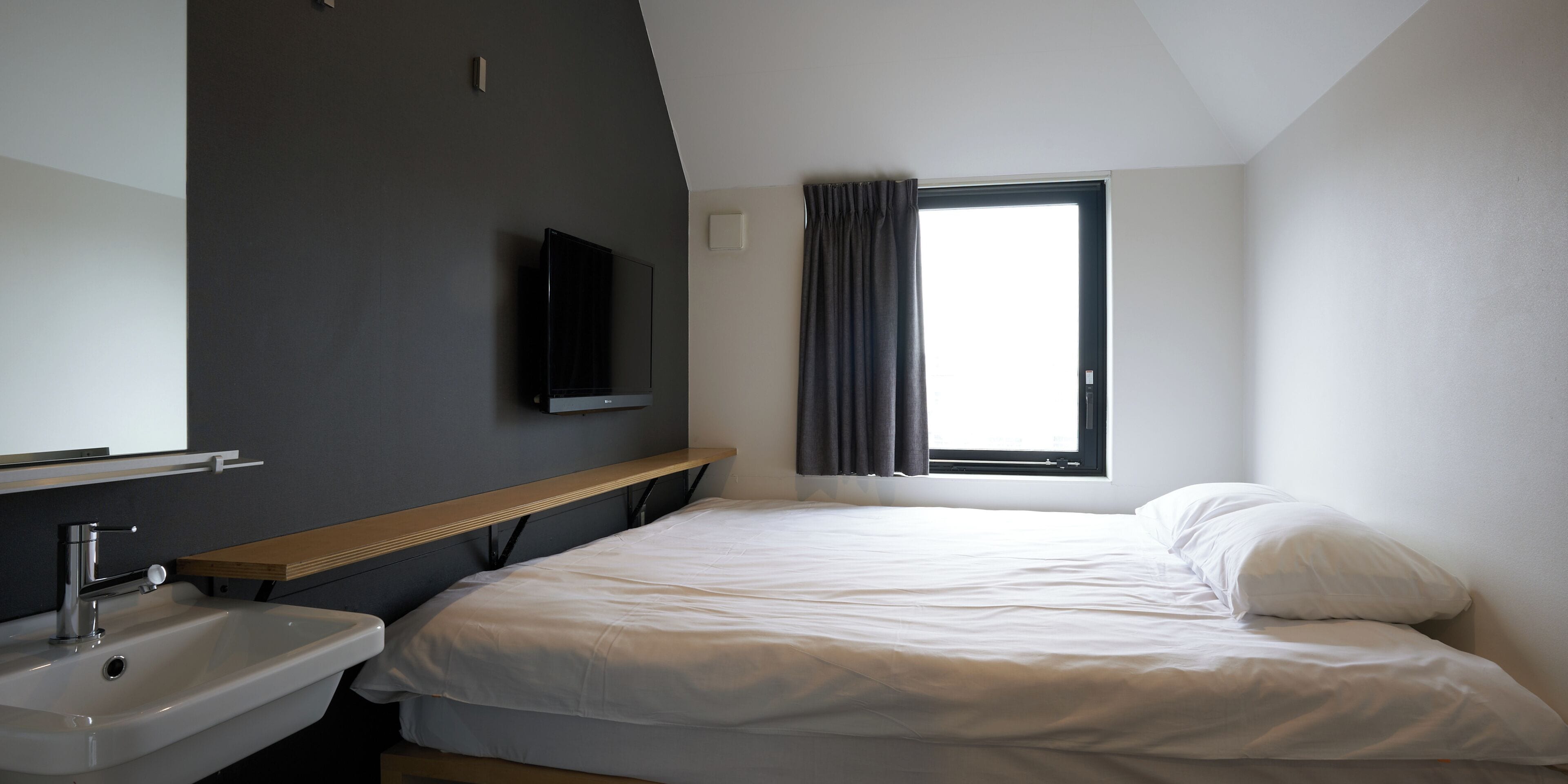 A bedroom with a bed, a bedside table, a mirror, a window with curtains, and a wall-mounted TV.
