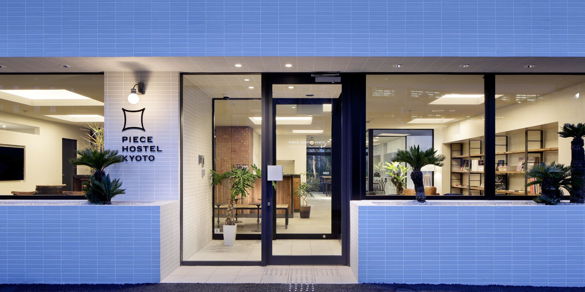 Une auberge de jeunesse moderne, au design épuré, dotée d’un espace d’accueil et d’un intérieur visible, aménagé avec des sièges et des plantes.