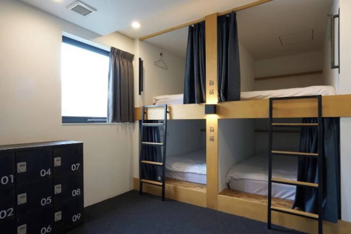 A dormitory room with bunk beds, a window with curtains, a wardrobe, and a desk with a chair.
