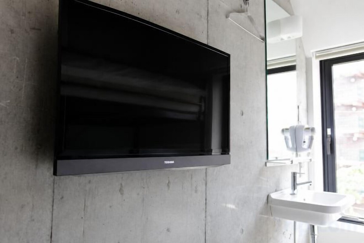 A flat-screen TV mounted on a concrete wall, a white sink, and a window with a view.