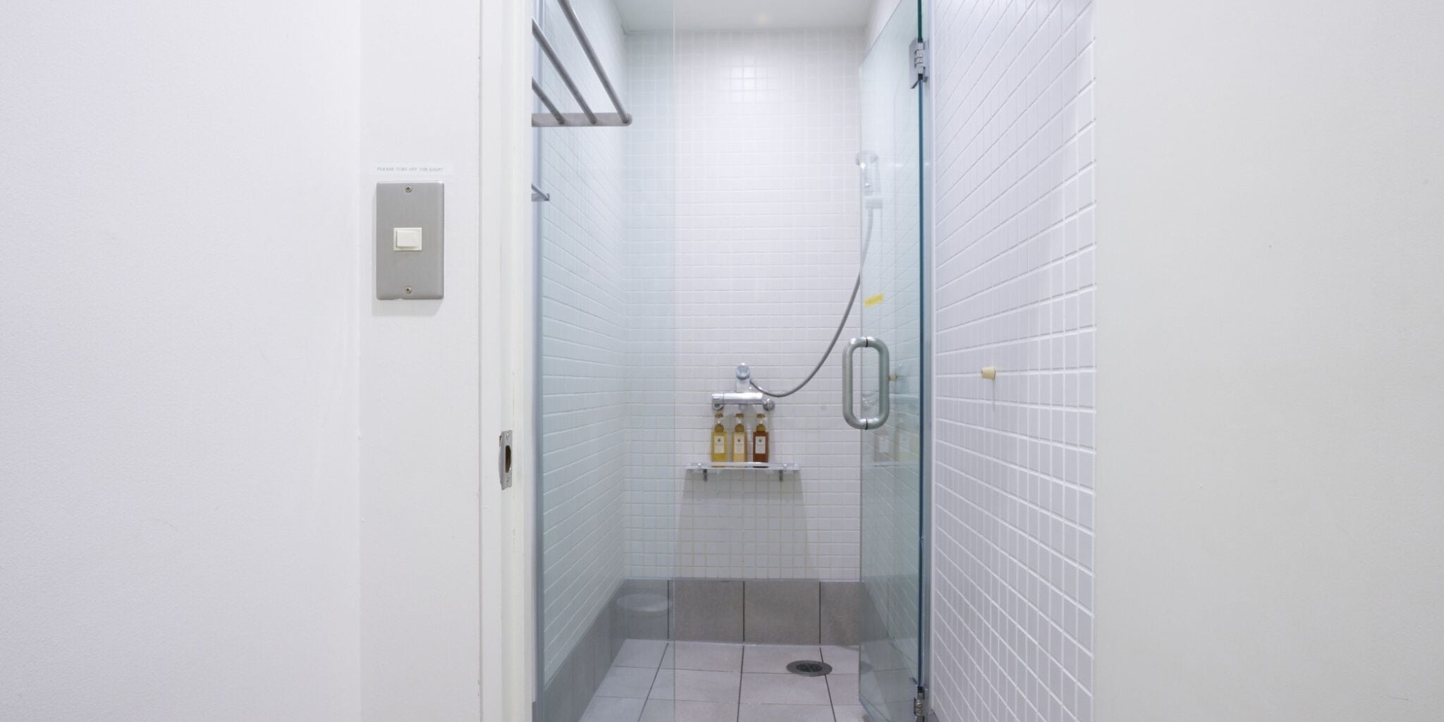 A narrow, tiled shower stall with a glass door, a towel rack with bottles, and a light switch on the wall.