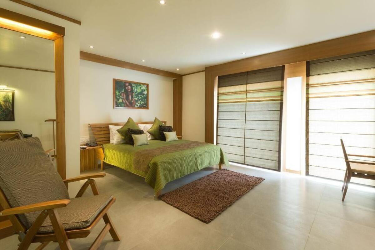 A bedroom with a bed, two chairs, a bedside table, a painting on the wall, and a large window with blinds.