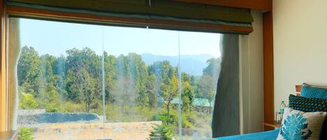 A bedroom with a large window, a bed with blue bedding, and a view of a natural landscape.