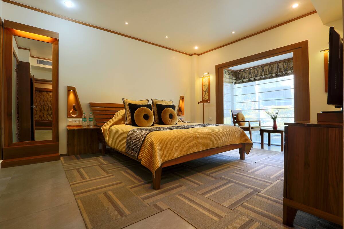 A bedroom with a large bed, a wooden headboard, a nightstand, a bedside table, a mirror, a window with curtains, and a television on a wooden stand.