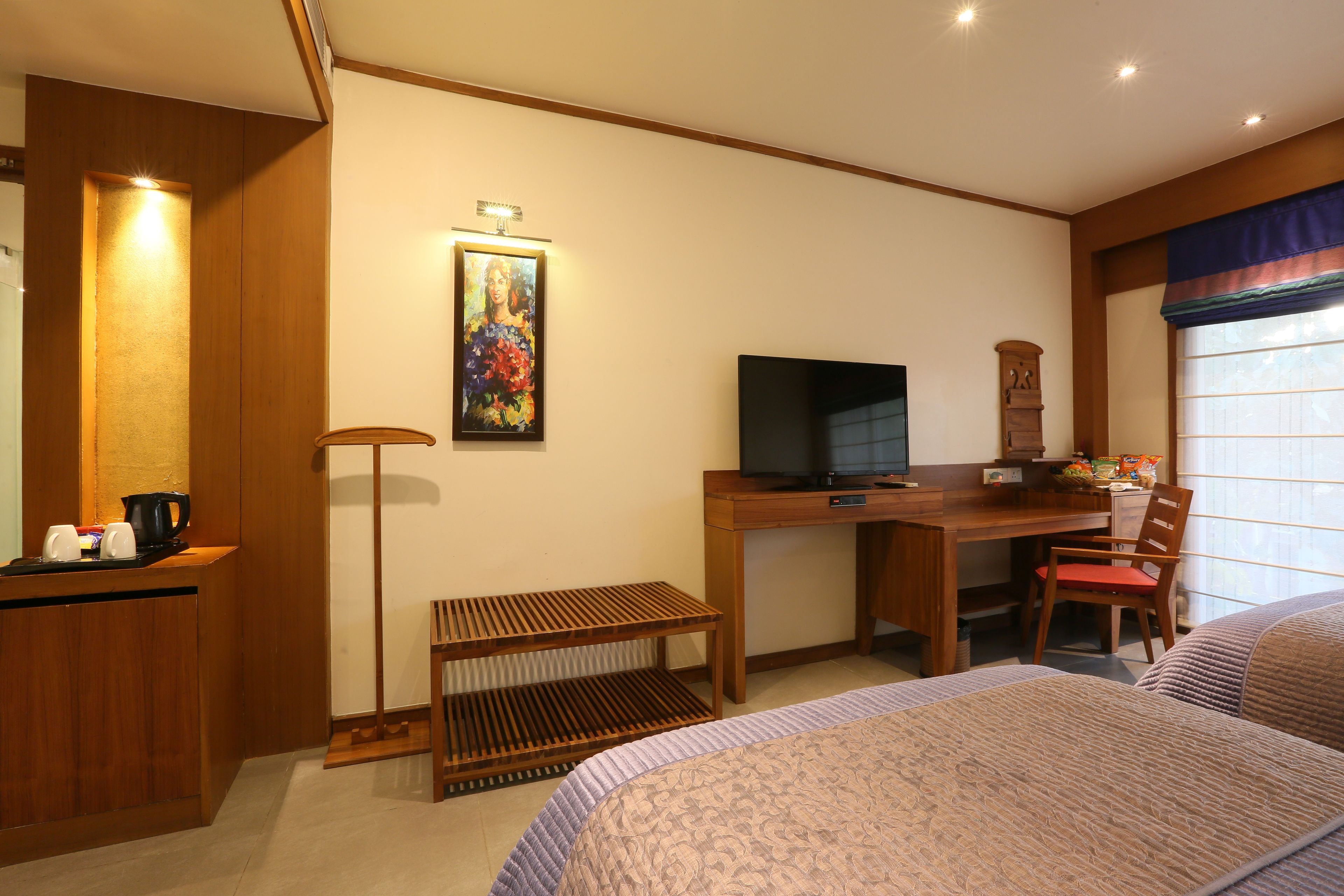 A hotel room with a bed, a desk with a TV, a chair, a small table with a kettle, and a painting on the wall.