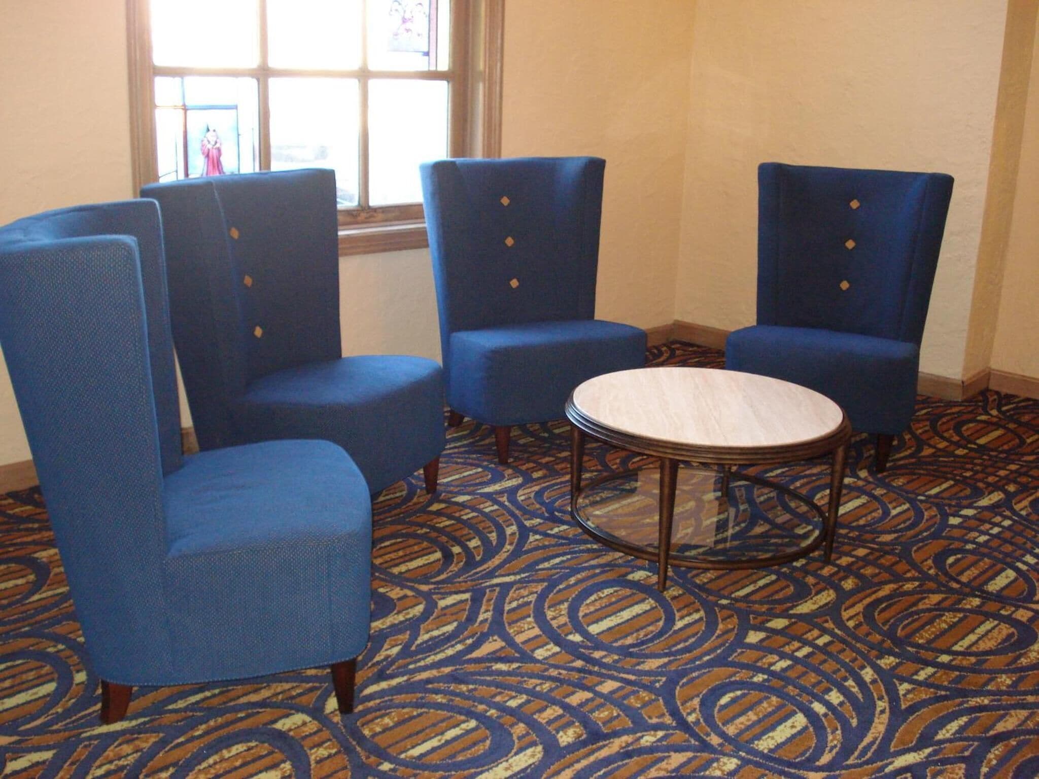 A room with four blue armchairs and a round table. The room has patterned carpet and a window with a view.