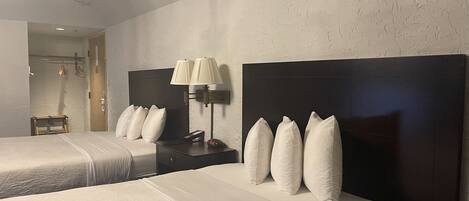 A hotel room with two beds, each with a dark headboard and white bedding. There is a nightstand with a lamp and a clock between the beds.