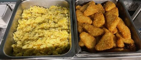 Two metal trays of food, one with scrambled eggs and the other with breaded chicken tenders.