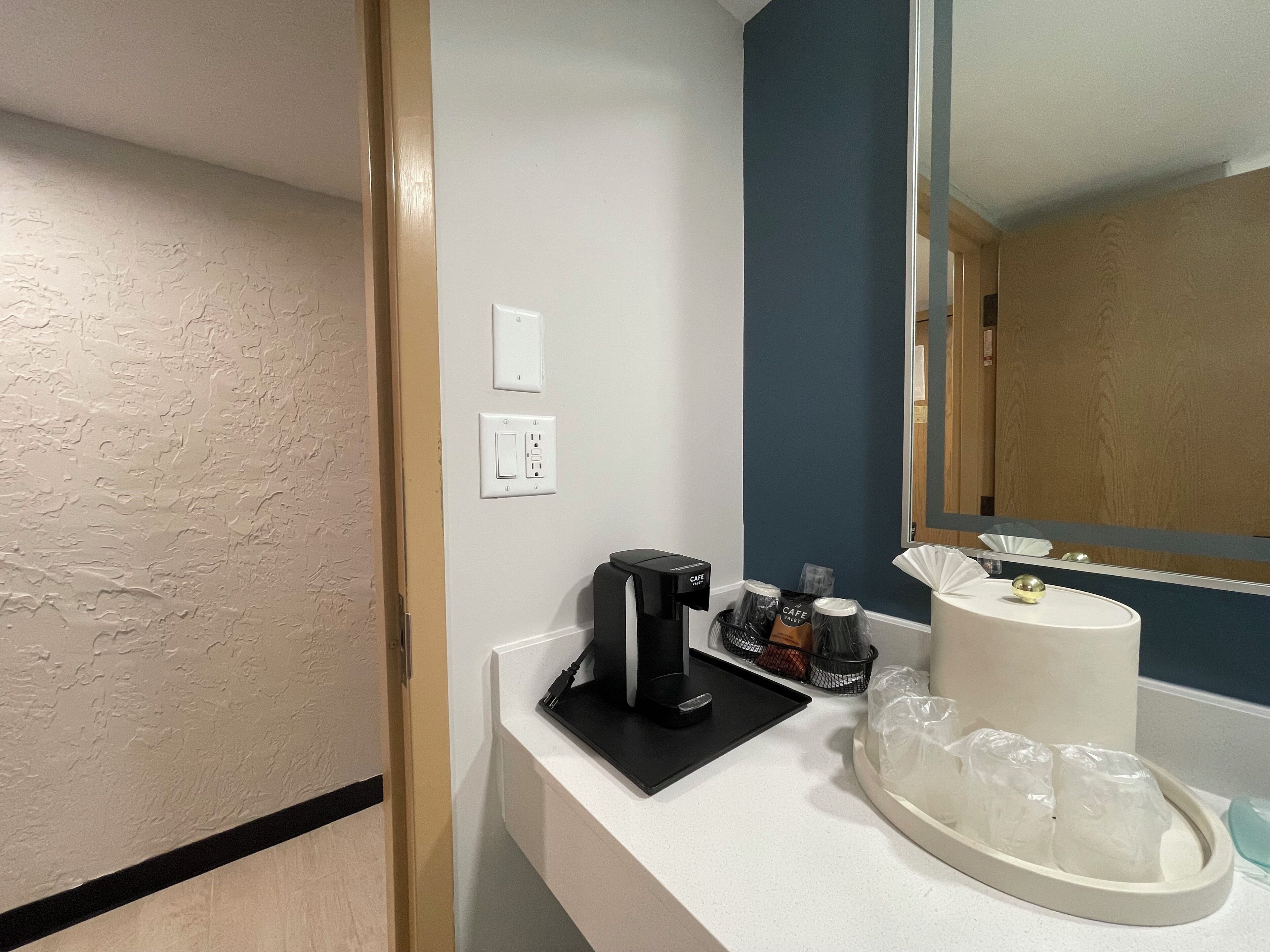 Habitación de hotel con un mostrador blanco, una cafetera, una bandeja con vasos desechables y un espejo que refleja la habitación.