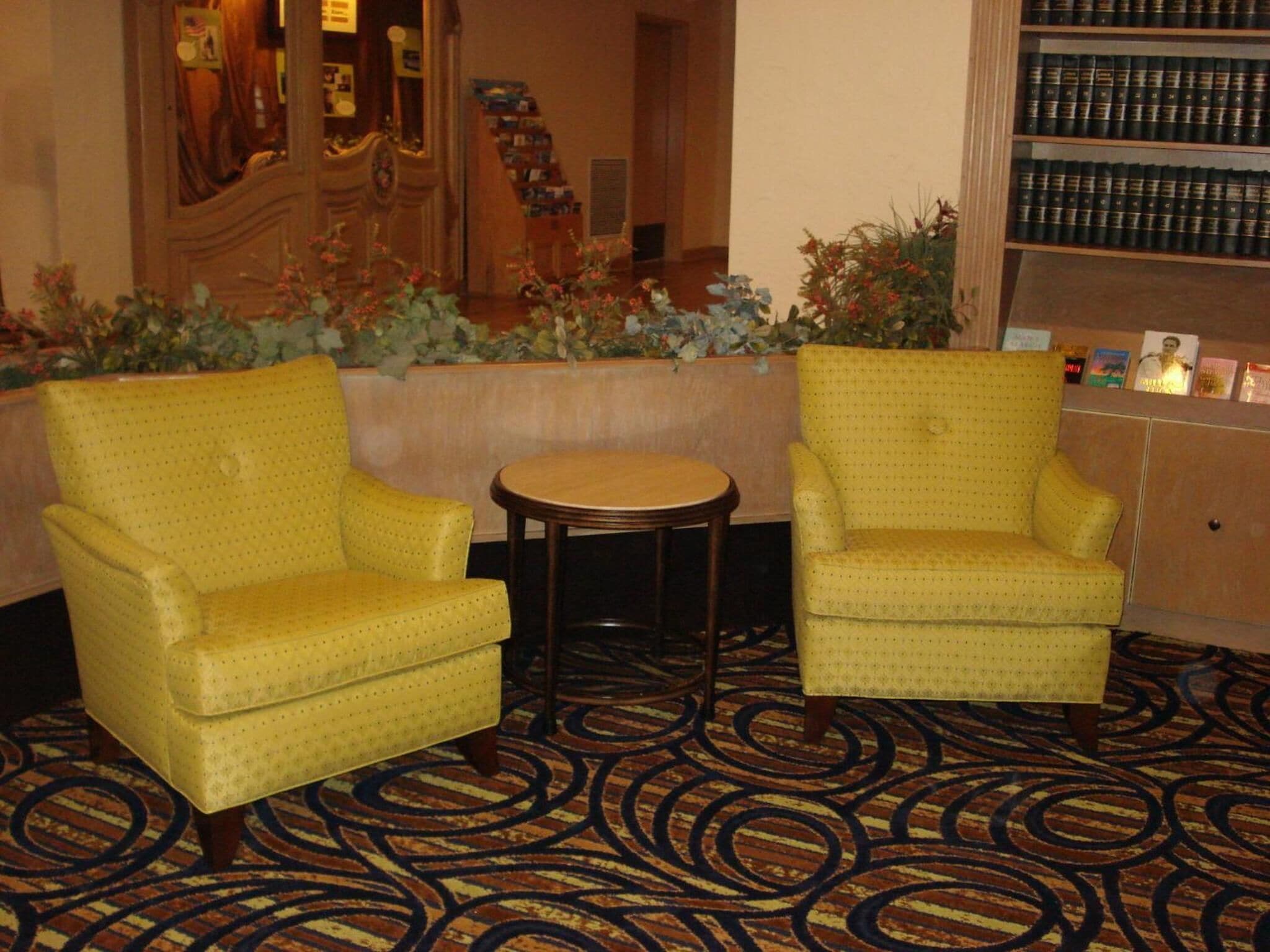 A hotel lobby with two yellow armchairs, a small round table, and a bookshelf filled with books.