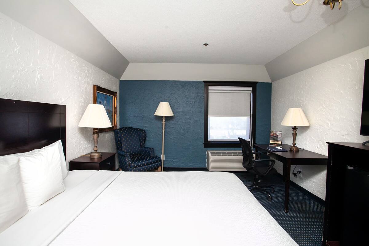 Hotellihuone, jossa on sänky, kaksi yöpöytävalaisinta, työpöytä tuolilla, sininen nojatuoli ja suuri ikkuna.