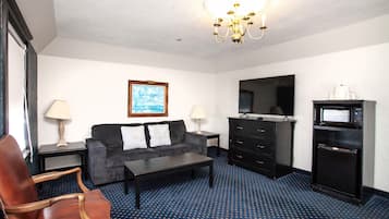 A hotel room with a sofa, armchair, coffee table, dresser, television, microwave, and a chandelier.