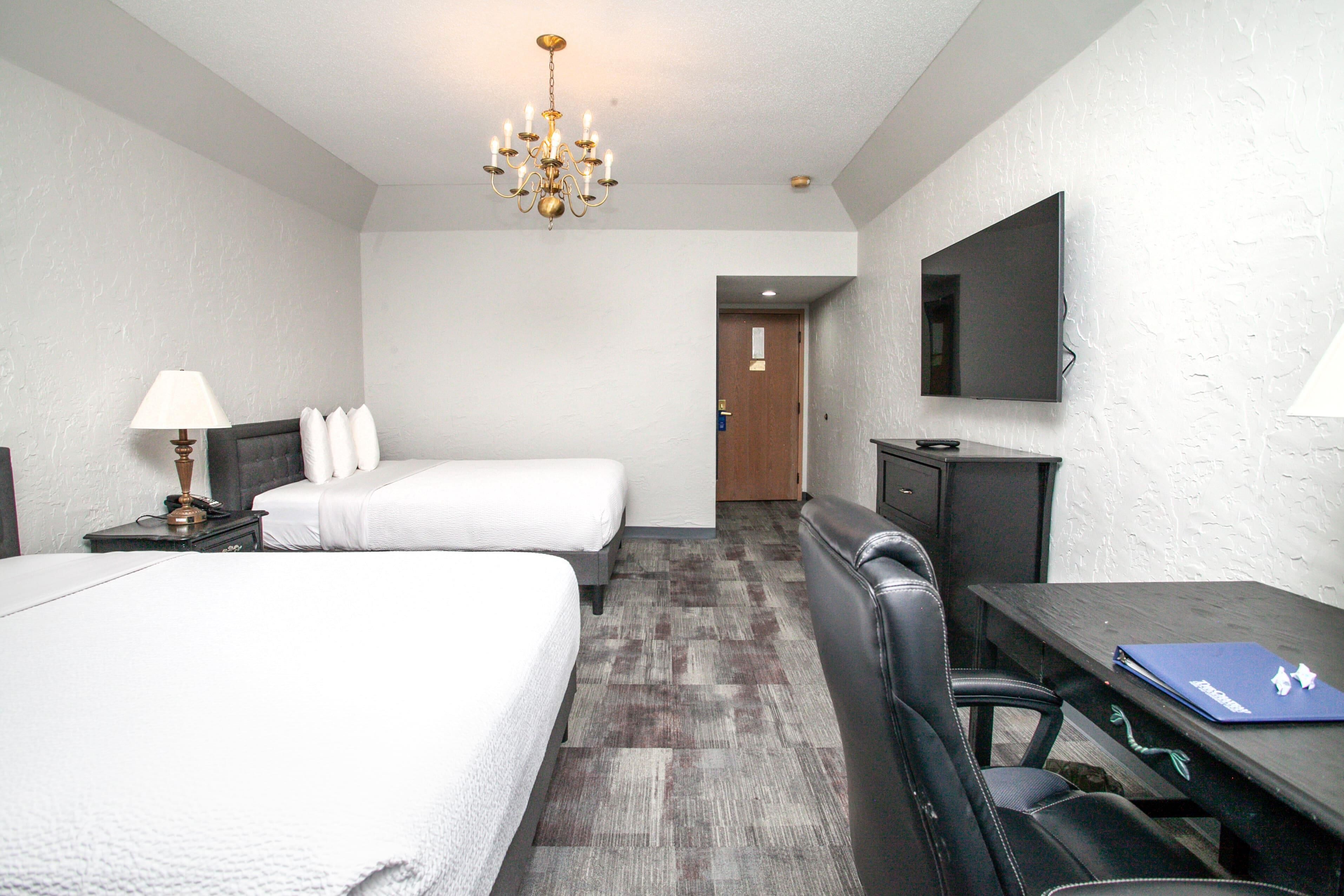 A hotel room with two beds, a desk, a chair, a TV, and a chandelier.