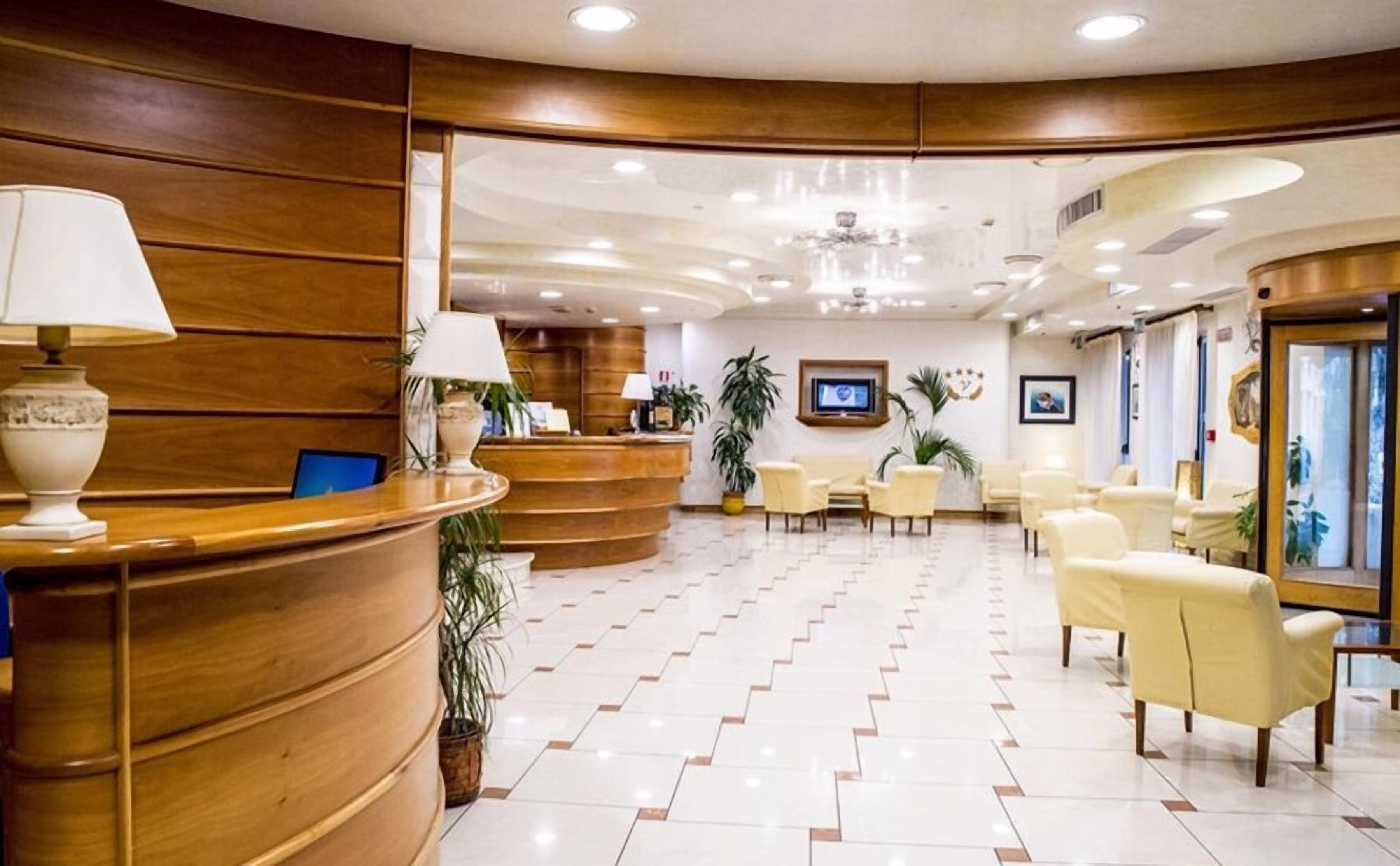 Zona de recepción de un hotel con un mostrador curvo, varias sillas, una pantalla de televisión y plantas en macetas.