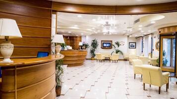 Ein Hotel-Empfangsbereich mit einer gebogenen Rezeption, einigen Stühlen, einem Fernsehbildschirm und Topfpflanzen.