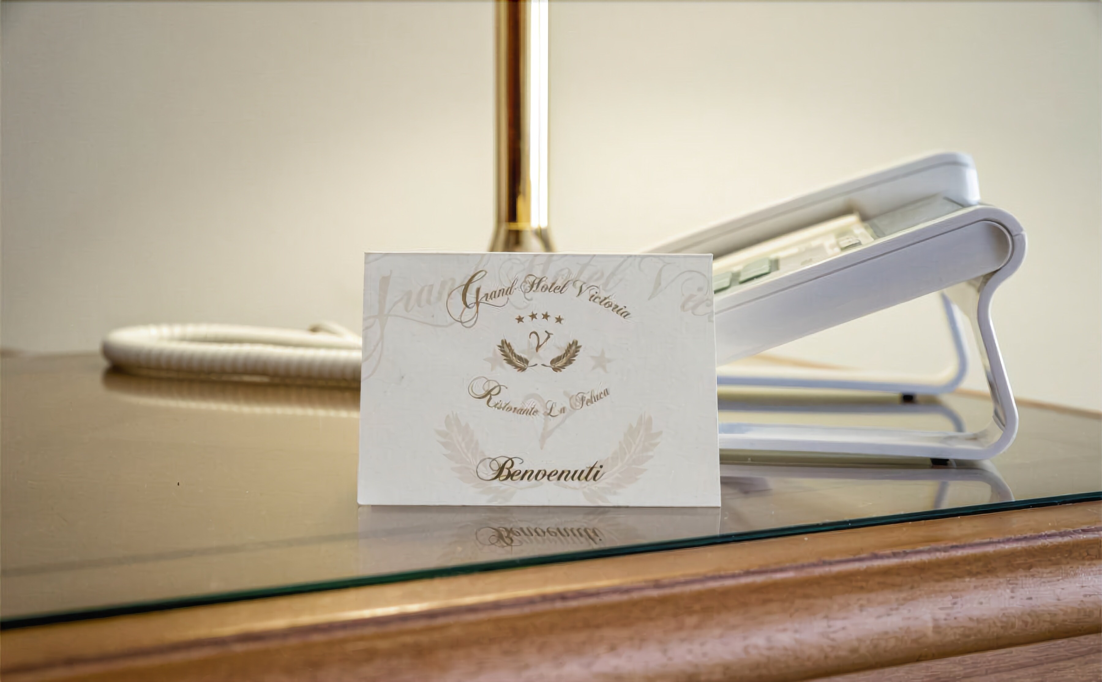 A hotel room with a glass table displaying a card that reads "Grand Hotel Victoria Restaurante La Feluca Benuenuti."