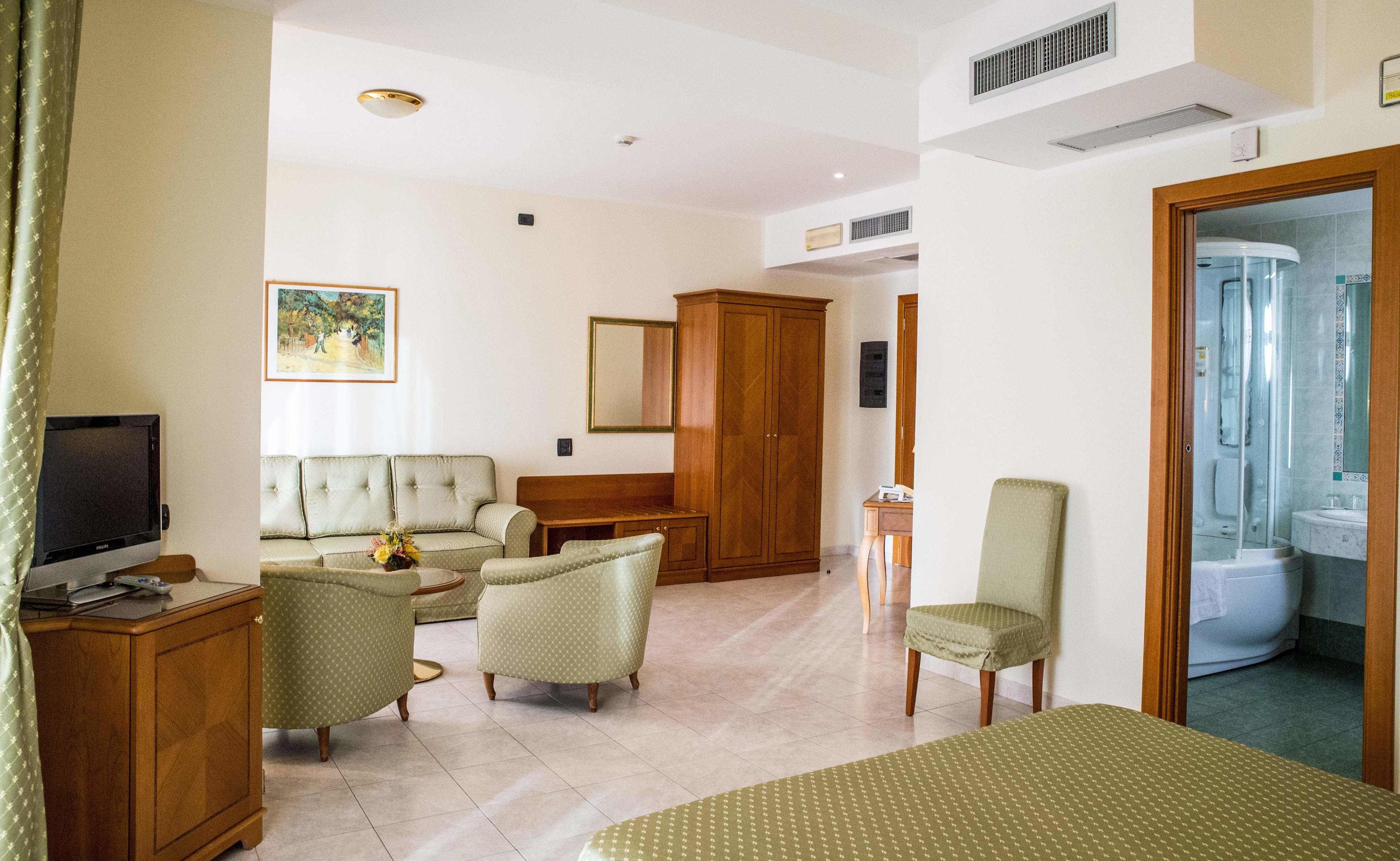 Habitación de hotel con sofá, sillones, televisor, armario, ropero de madera y baño con ducha.