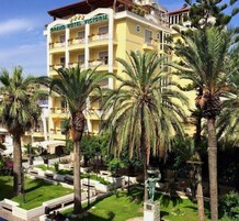 Un hotel de varios pisos con balcones, rodeado de palmeras y un jardín cuidado.
