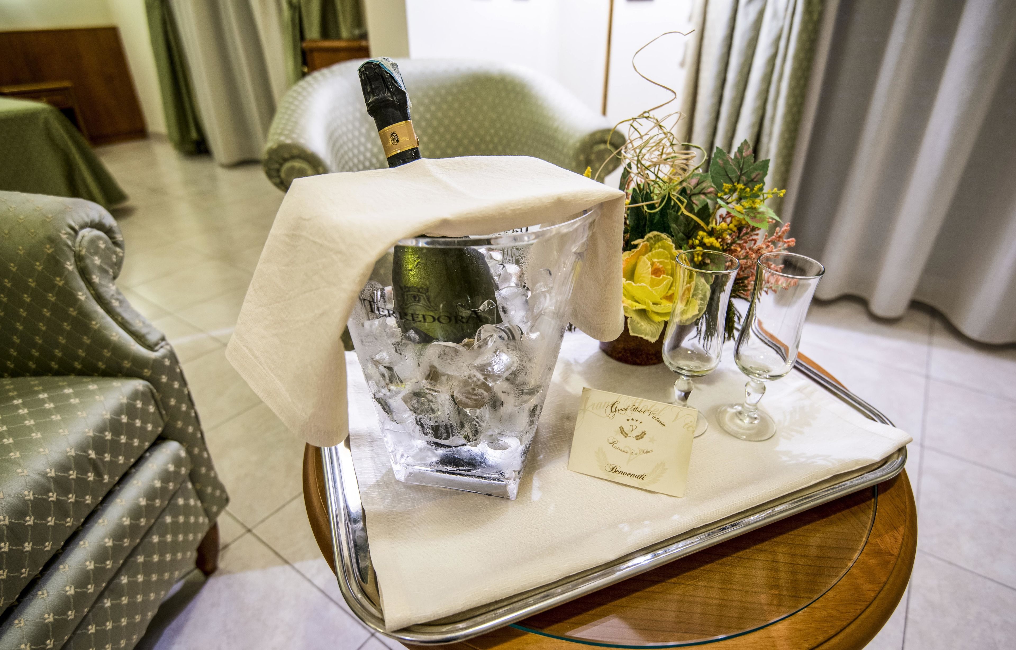 A room with a bed, a green patterned armchair, a glass table with a bottle of champagne, a vase with flowers, and two wine glasses.