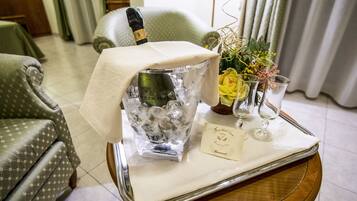 Una stanza con un letto, una poltrona verde con motivo decorativo, un tavolino in vetro con una bottiglia di champagne, un vaso con fiori e due bicchieri da vino.