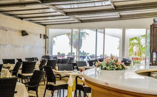 Un restaurante con terraza, amplios ventanales y vistas a palmeras.