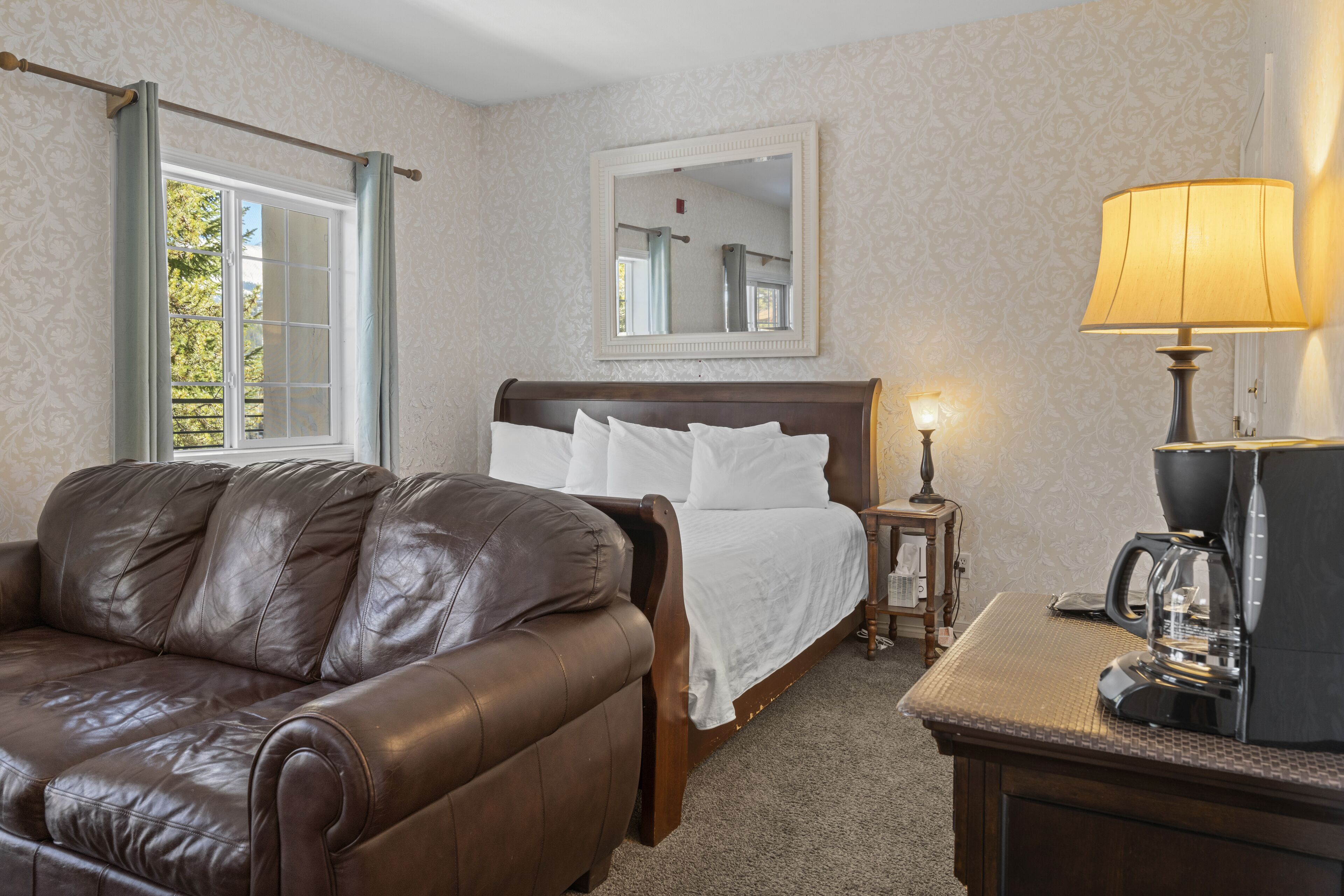 A hotel room with a leather sofa, a bed with white linens, a bedside lamp, and a coffee maker on a wooden table.