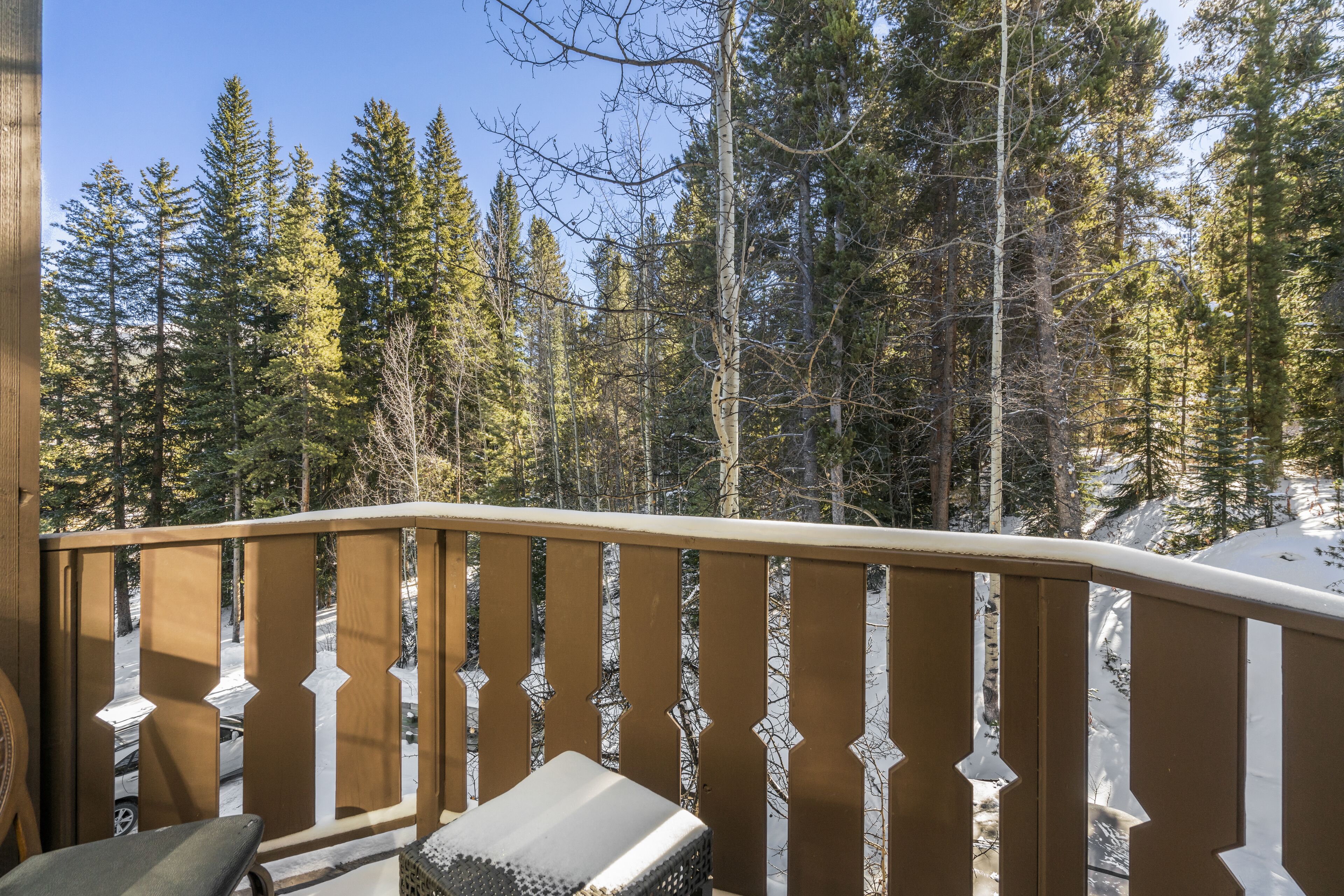 A wooden balcony with a snow-covered bench and railing, overlooking a forested area with tall trees.