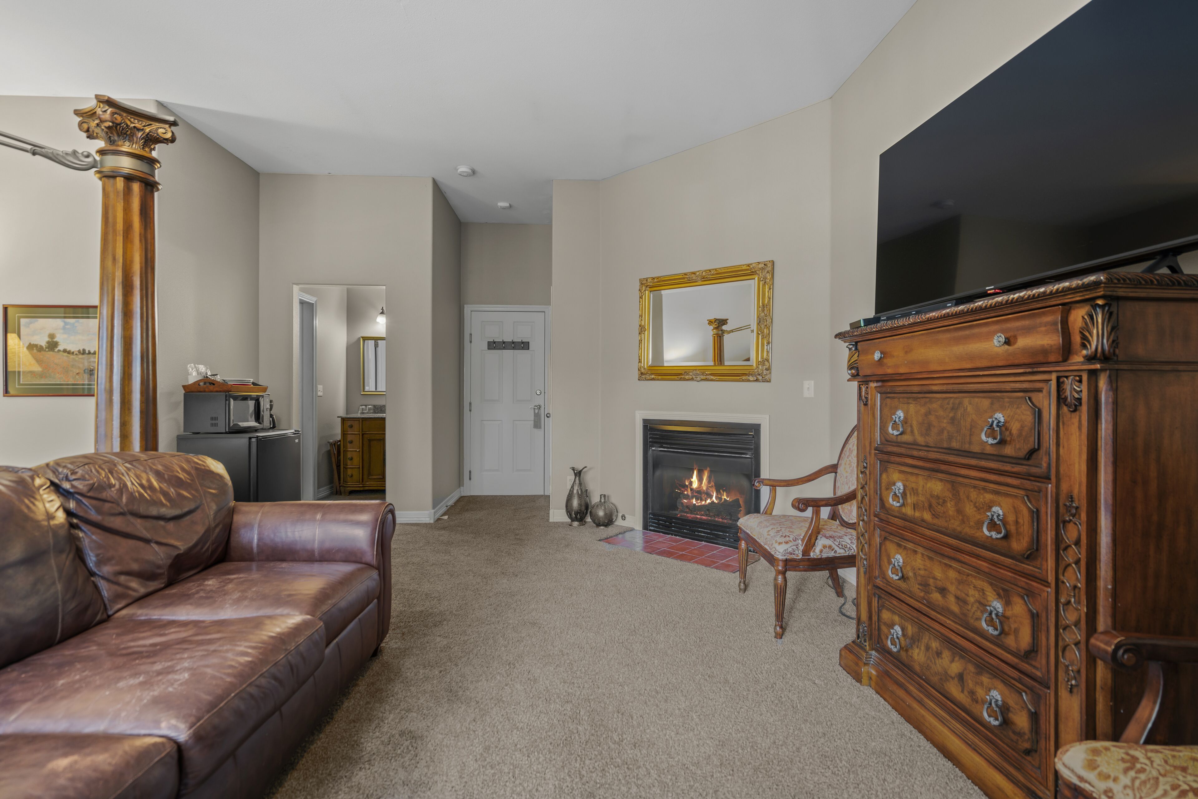 A living room with a leather couch, a wooden dresser, a television, a fireplace, and a door leading to another room.