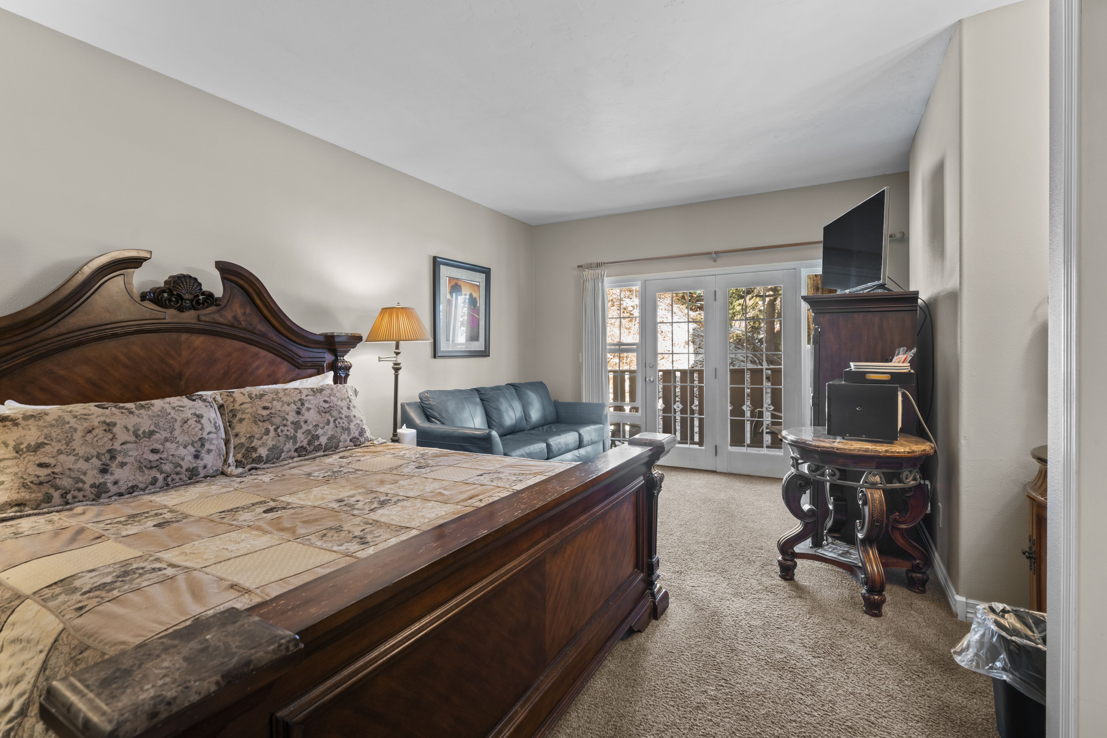 A bedroom with a large bed, a dark leather sofa, a wooden side table with a laptop, and a door leading to a balcony.