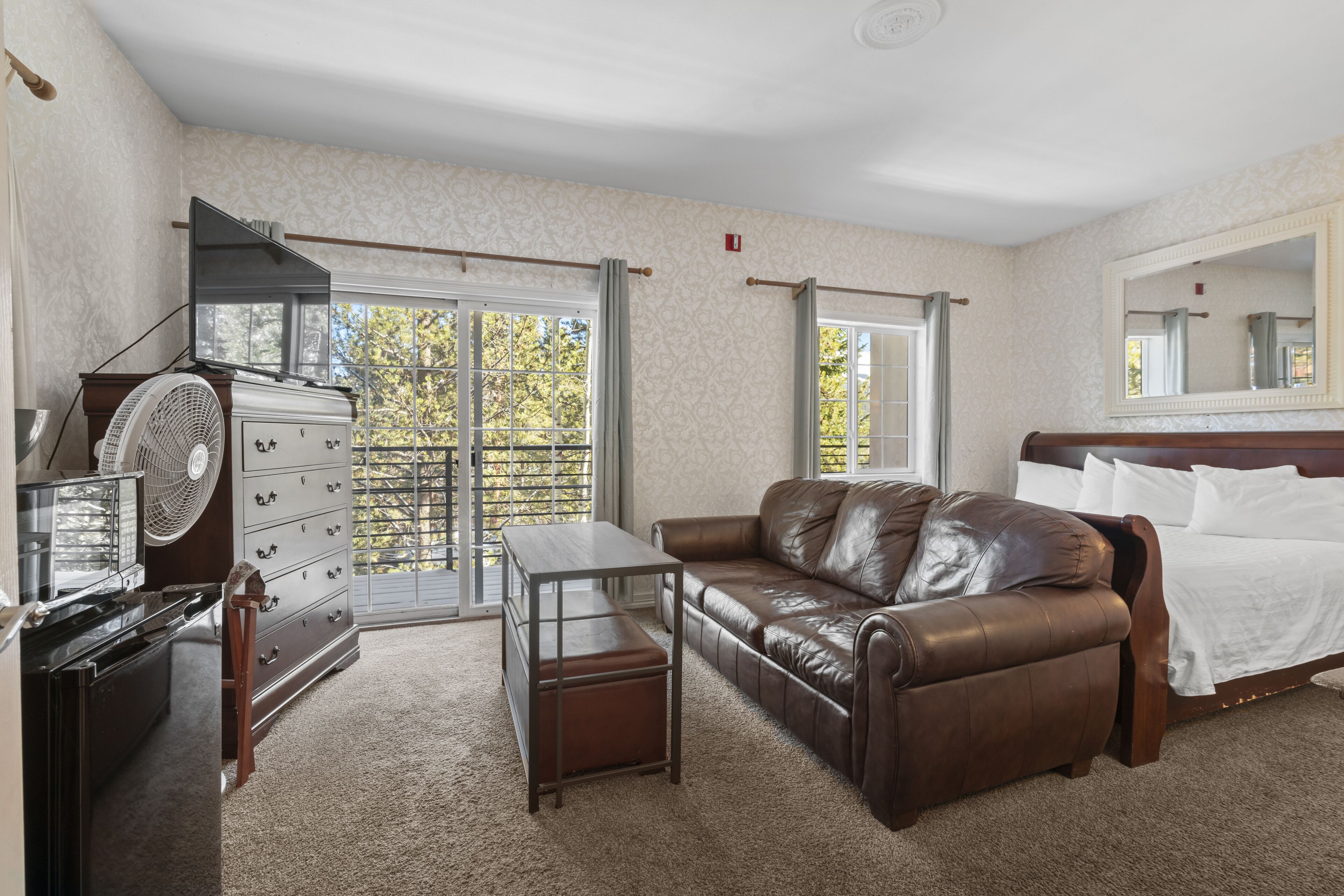 A room with a brown leather sofa, a wooden dresser, a small table, and a fan. There is a window with a view of trees and a door leading outside.