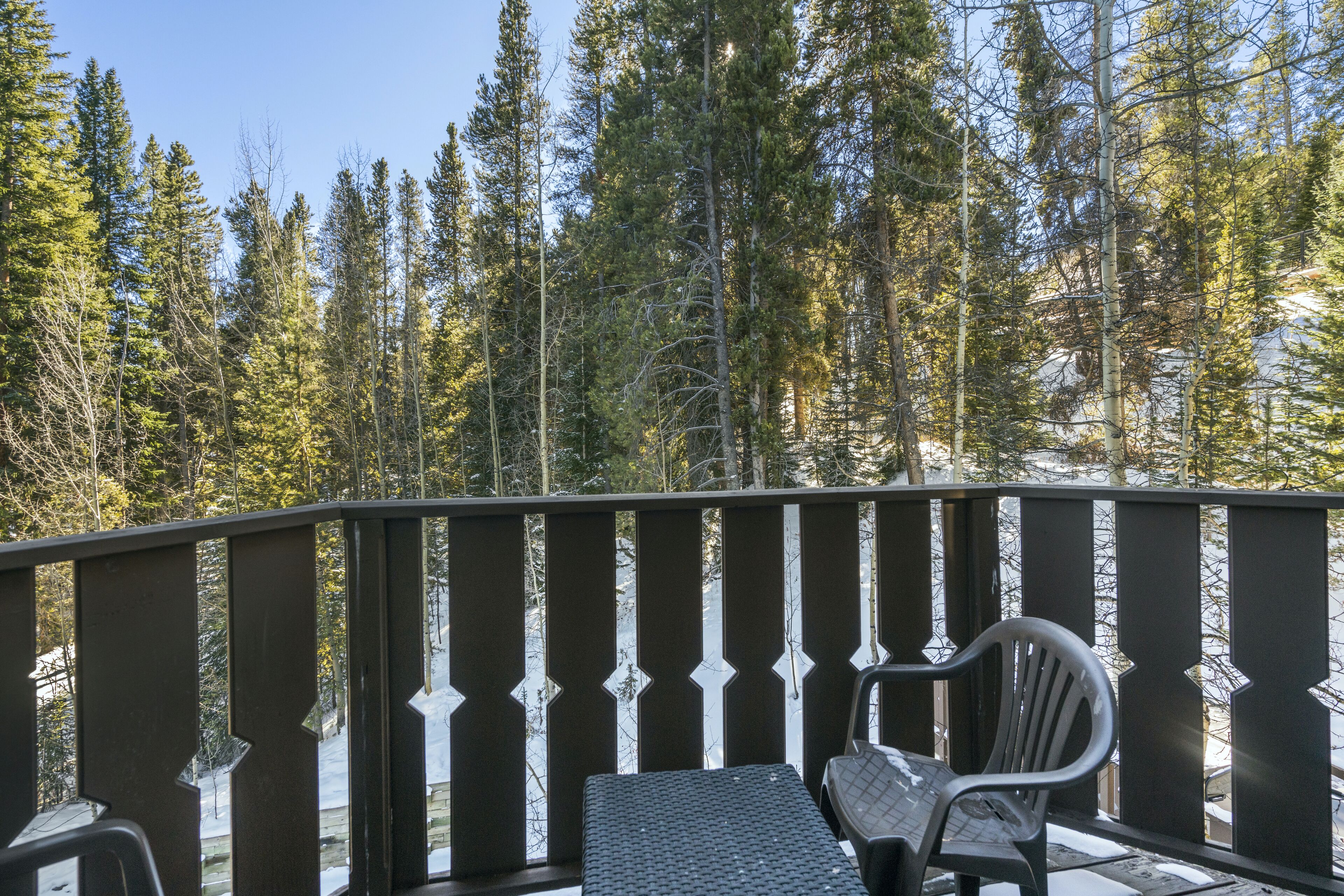 A deck with a metal railing, a table, and two chairs, overlooking a forested area with snow on the ground.
