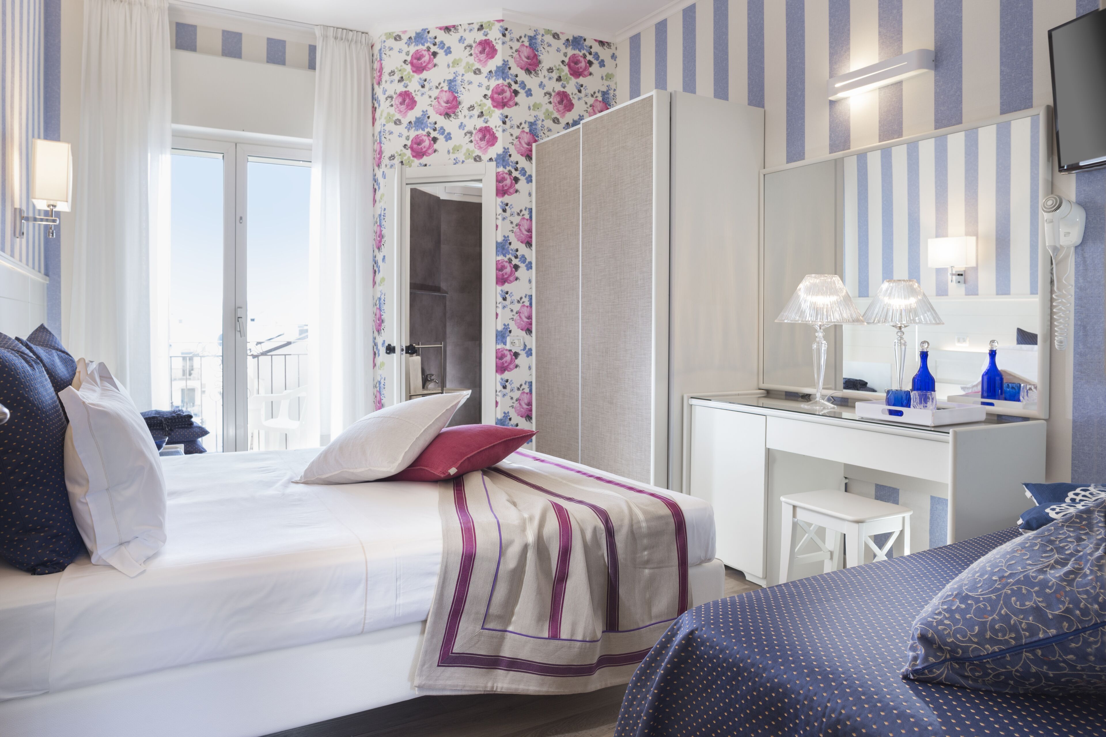 A hotel room with a bed, a desk, a mirror, and a view of the outside.