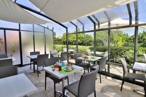 Un patio al aire libre con mesas y sillas, amplias ventanas de vidrio y vistas al follaje.