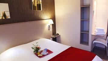 A hotel room with a bed, a bedside table with a lamp, a small desk with a chair, and a window.