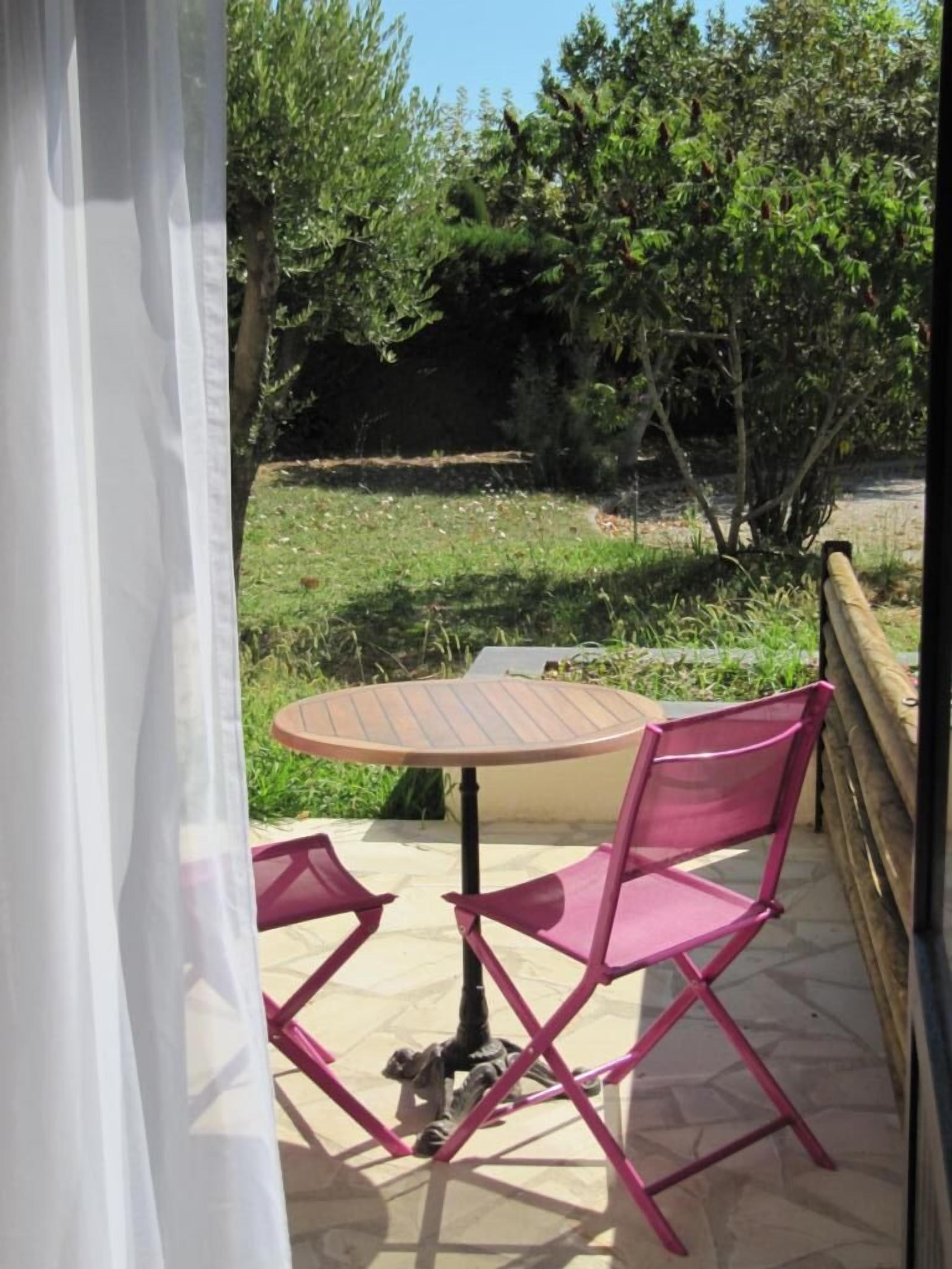 Eine Terrasse mit einem runden Tisch und zwei rosa Stühlen, umgeben von Grünpflanzen und einem Holzzaun.