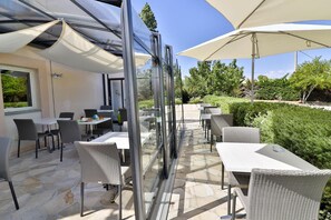 An outdoor patio with glass walls, wicker furniture, and large umbrellas. - Hôtel Le Coudon (La Farlede)